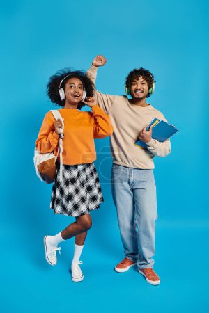 An interracial couple of students in casual attire standing closely together against a blue backdrop in a studio.