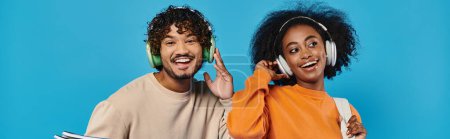 Téléchargez les photos : Un homme et une femme, étudiants interraciaux, se tiennent ensemble avec des écouteurs dans un studio sur fond bleu. - en image libre de droit