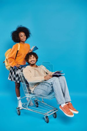 indian man sits on a shopping cart while a woman stands beside her, posing for a fun and quirky urban portrait.