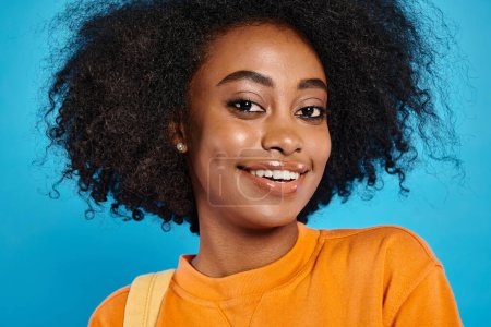 Una chica universitaria afroamericana con un impresionante peinado afro sonríe brillantemente con atuendo casual en un fondo de estudio azul.