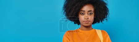 Photo for A stylish African American woman in casual attire stands confidently with her voluminous afro hair, in front of a vibrant blue backdrop. - Royalty Free Image