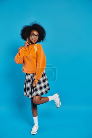 Une étudiante afro-américaine élégante se tient avec confiance dans un pull orange vif et une jupe à carreaux tendance sur fond bleu.