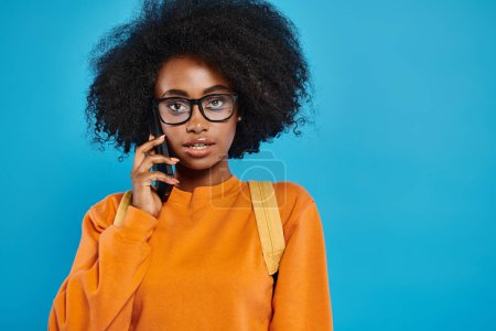 An African American college girl with glasses chats on a cell phone against a blue backdrop in a studio setting.