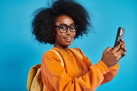An African American college girl in casual attire, wearing glasses, holding a cell phone in a studio with a blue backdrop.