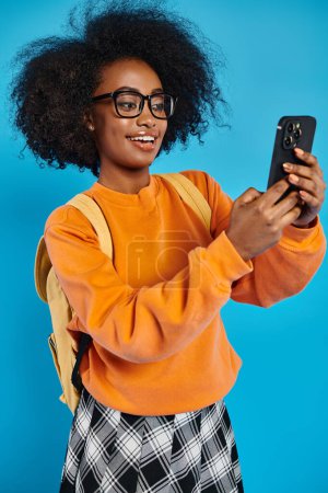 A young African American woman in casual attire takes a selfie with her cell phone while carrying a backpack, set against a blue backdrop in a studio.