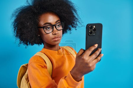 An African American college girl in casual attire, wearing glasses, taking a selfie with her cell phone on a blue backdrop in a studio.