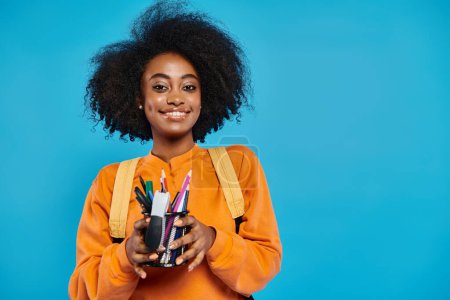 Ein afroamerikanisches College-Mädchen in lässiger Kleidung hält vor blauem Hintergrund eine mit Stiften und Bleistiften gefüllte Tasse.