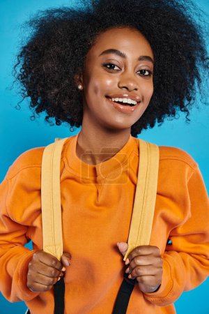 A joyful African American college girl with an afro hairstyle smiles while holding a pair of suspenders, standing in casual attire against a blue backdrop in a studio.