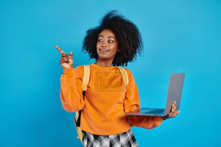 Photo for An African American college girl in casual attire holding a laptop, pointing to the side in a studio with a blue backdrop. - Royalty Free Image