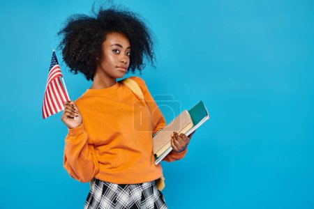 Una joven universitaria afroamericana de pie con orgullo, sosteniendo un libro y una bandera americana.