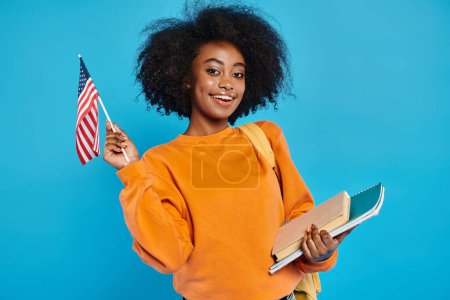 An African American college girl proudly holds a book and an American flag in a studio setting.