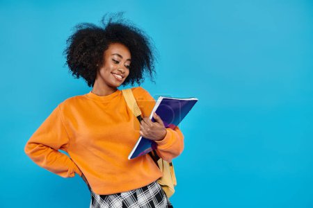 Colegiala afroamericana con camisa naranja sosteniendo un libro, exudando conocimiento e inspiración en un ambiente de estudio.