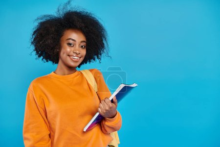 Ein junges afroamerikanisches College-Mädchen steht in lässiger Kleidung, hält ein Buch in der Hand und lächelt freundlich in die Kamera vor blauem Hintergrund..