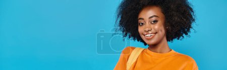 Una chica universitaria con un gran afro sonríe calurosamente a la cámara contra un telón de fondo azul en un entorno de estudio.
