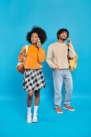 An interracial couple of students with casual attire stand together against a blue backdrop in a studio.