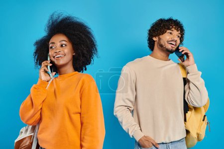 Photo for A man and a woman, interracial students, standing together in casual attire, engaged in conversation on cell phones. Blue backdrop in studio. - Royalty Free Image