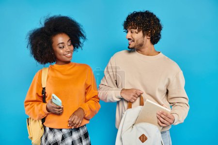 Photo for A man and a woman, interracial students, stand together in casual attire against a blue backdrop in a studio. - Royalty Free Image
