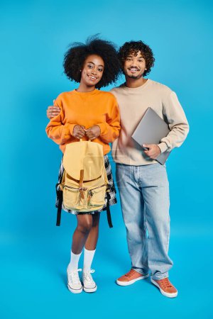 An interracial couple of students stand together in casual attire against a blue backdrop in a studio.
