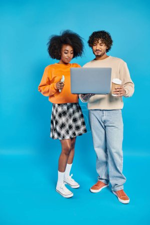 An interracial couple, standing together, holding a laptop against a blue backdrop.