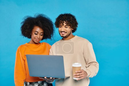 Téléchargez les photos : Un homme et une femme, tenant un ordinateur portable et une tasse de café, debout ensemble dans un studio avec une toile de fond bleue. - en image libre de droit