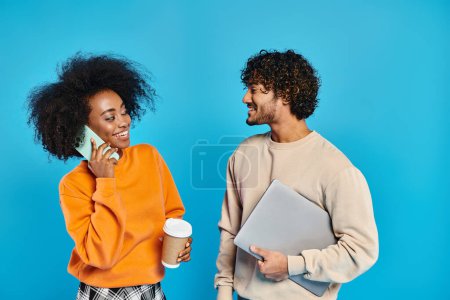 Téléchargez les photos : Un couple interracial d'étudiants debout ensemble en tenue décontractée sur fond bleu, à l'aide d'appareils - en image libre de droit