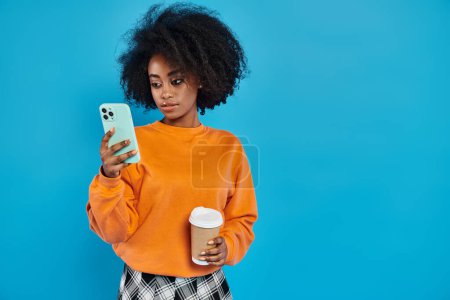 black woman stands holding a cup of coffee and a cell phone, showcasing the art of multitasking in a modern world.