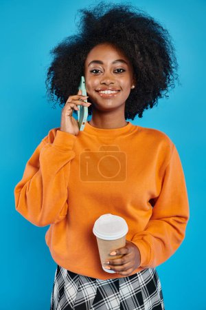Una mujer con estilo de diferentes etnias sosteniendo una taza de café mientras habla en un teléfono celular contra un telón de fondo azul.