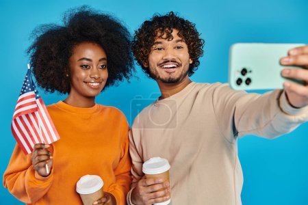 An interracial man and woman in casual attire take a selfie with American flag against a blue backdrop in a studio.