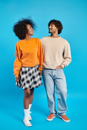 An interracial couple of students in casual attire stands next to each other against a blue backdrop in a studio setting.