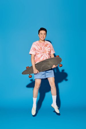 Téléchargez les photos : Homme élégant en tenue tendance tenant skateboard dans les mains sur fond bleu. - en image libre de droit
