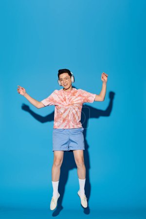 Téléchargez les photos : Un jeune homme à la mode en tenue tendance saute joyeusement dans les airs avec les mains levées sur un fond bleu. - en image libre de droit