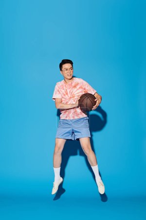 Un jeune homme élégant pose avec un ballon de basket sur fond bleu vif, respirant la confiance et l'athlétisme.