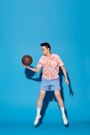 A stylish young man in trendy attire confidently holds a basketball in his right hand against a vibrant blue backdrop.