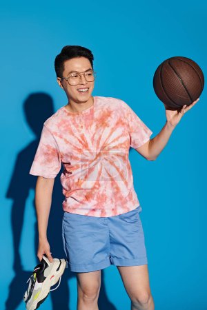 Un joven elegante con un atuendo de moda sostiene enérgicamente una pelota de baloncesto en su mano contra un telón de fondo azul.