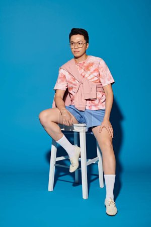 A stylish young man in trendy attire confidently sits atop a white stool against a vibrant blue backdrop.