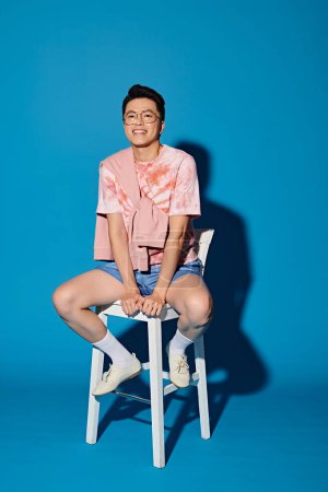 A stylish young man in trendy attire poses confidently while sitting on top of a white chair against a blue backdrop.