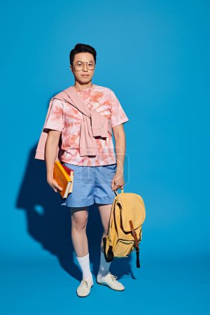 Stylischer junger Mann in rosa Hemd und blauer Shorts, der energisch eine gelbe Tasche vor blauem Hintergrund hält.