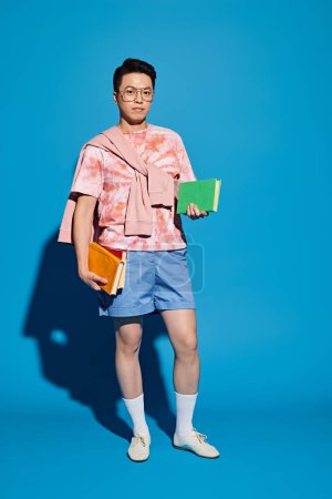 Un joven elegante con una camisa rosa y pantalones cortos azules sostiene un libro mientras posa con confianza sobre un telón de fondo azul.
