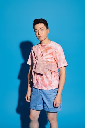 Ein stilvoller junger Mann in trendiger Kleidung posiert vor einer leuchtend blauen Wand und strahlt Zuversicht und Charme aus.