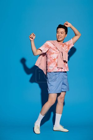 Un joven con estilo en una camisa rosa y pantalones cortos azules posa enérgicamente contra un telón de fondo azul, mostrando atuendo de moda.