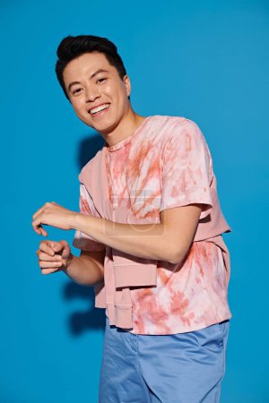 Stylischer junger Mann posiert energisch in rosa Hemd und blauer Hose vor leuchtend blauem Hintergrund.