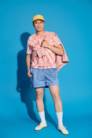 Un joven, vestido con estilo con una camisa rosa y pantalones cortos azules, posa enérgicamente sobre un telón de fondo azul.