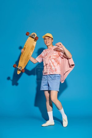 A stylish young man in trendy attire poses energetically, holding a skateboard, against a vibrant blue backdrop.