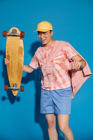 Junger Mann in schicker Kleidung hält selbstbewusst Skateboard vor leuchtend blauer Wand.