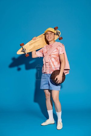 Ein stylischer junger Mann in trendiger Kleidung balanciert mühelos zwischen Skateboard und Ball vor leuchtend blauer Kulisse.