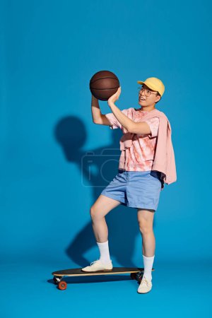 Homme élégant tenant un ballon de basket tout en équilibrant sur une planche à roulettes sur fond bleu.