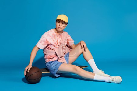Ein stylischer, gut aussehender junger Mann in trendiger Kleidung sitzt auf dem Boden und hält einen Basketball vor blauem Hintergrund.