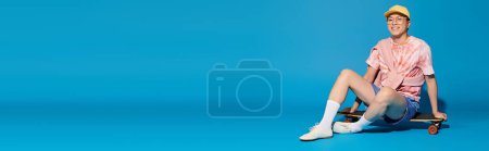 Photo for A stylish young man in trendy attire happily sits on a skateboard against a vibrant blue backdrop. - Royalty Free Image