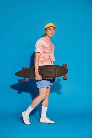 A stylish young man in trendy attire holds a skateboard on a vibrant blue background.