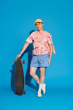 Ein stilvoller Mann in trendiger Kleidung hält energisch ein Skateboard vor leuchtend blauer Kulisse.
